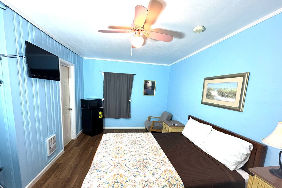 A bedroom with a bed, a ceiling fan, a television, a microwave, a small table, and a chair.