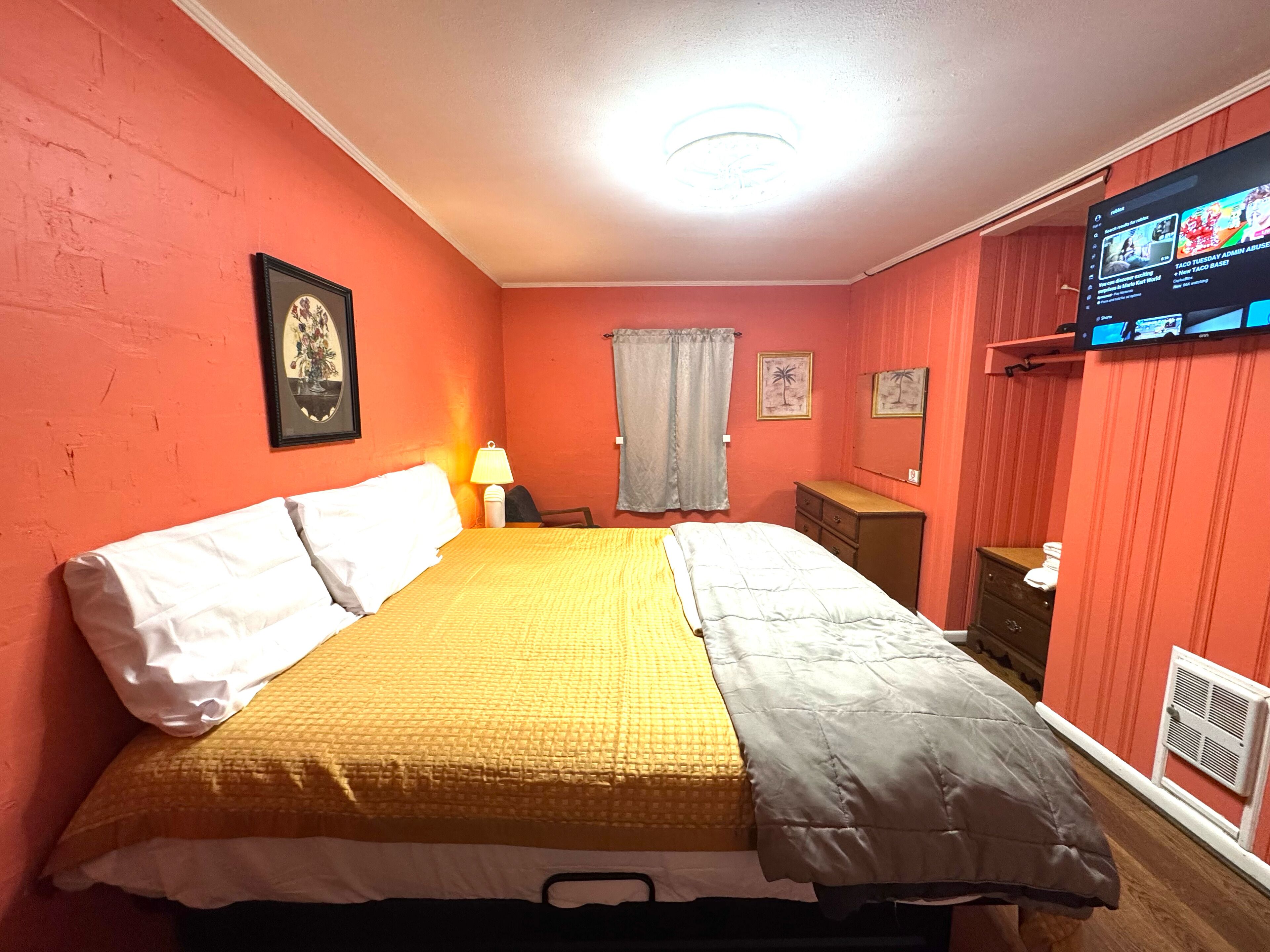 A bedroom with a bed, a nightstand, a lamp, a TV, and a window with curtains.