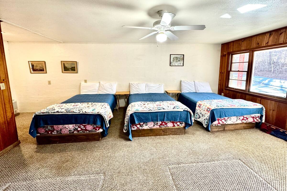 A room with three beds, a ceiling fan, and a window with a view of trees.