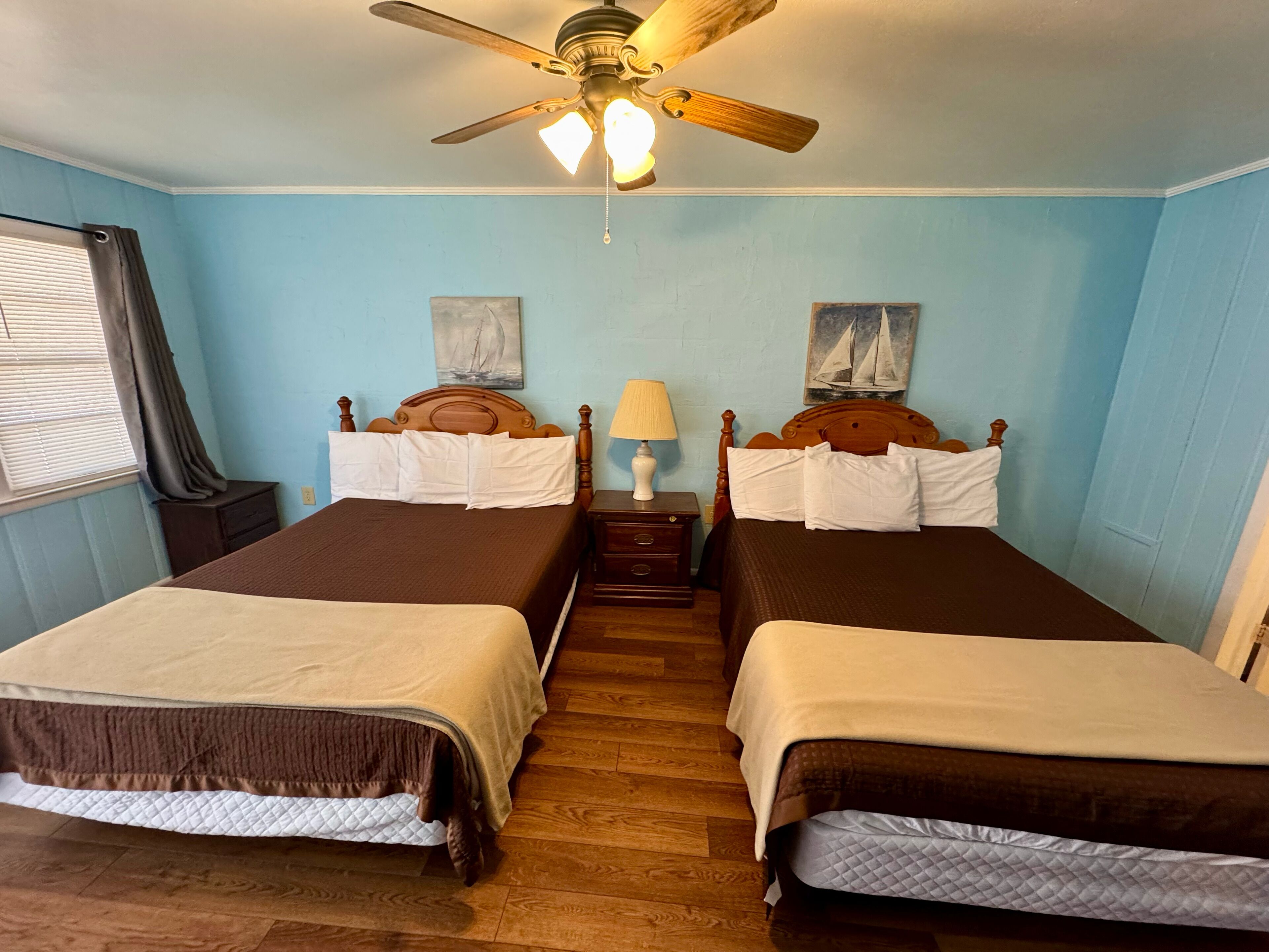 A room with two beds, wooden headboards, a ceiling fan, and two paintings on the walls.