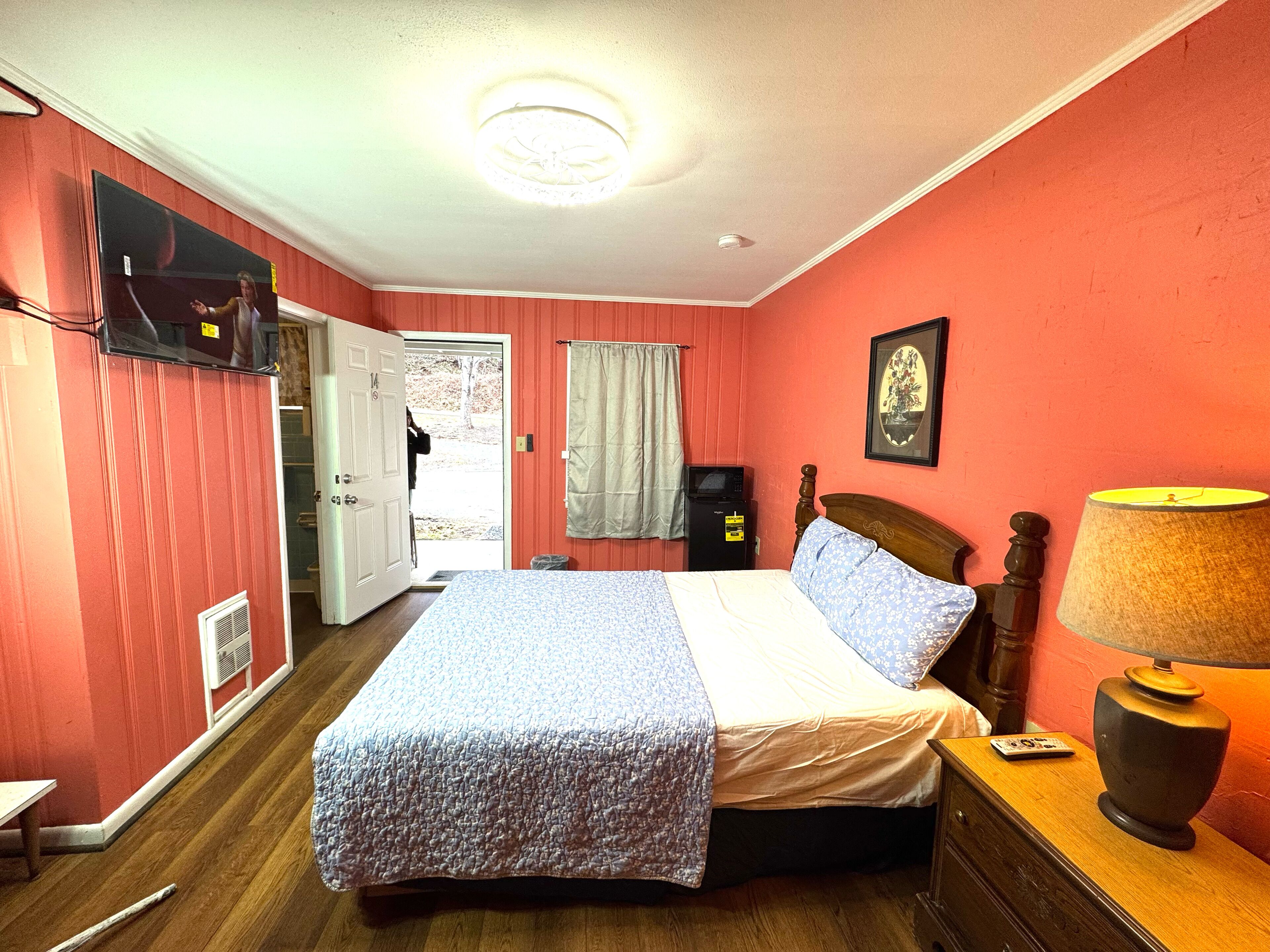 A bedroom with a bed, a bedside table with a lamp, a television on the wall, and a door leading outside.