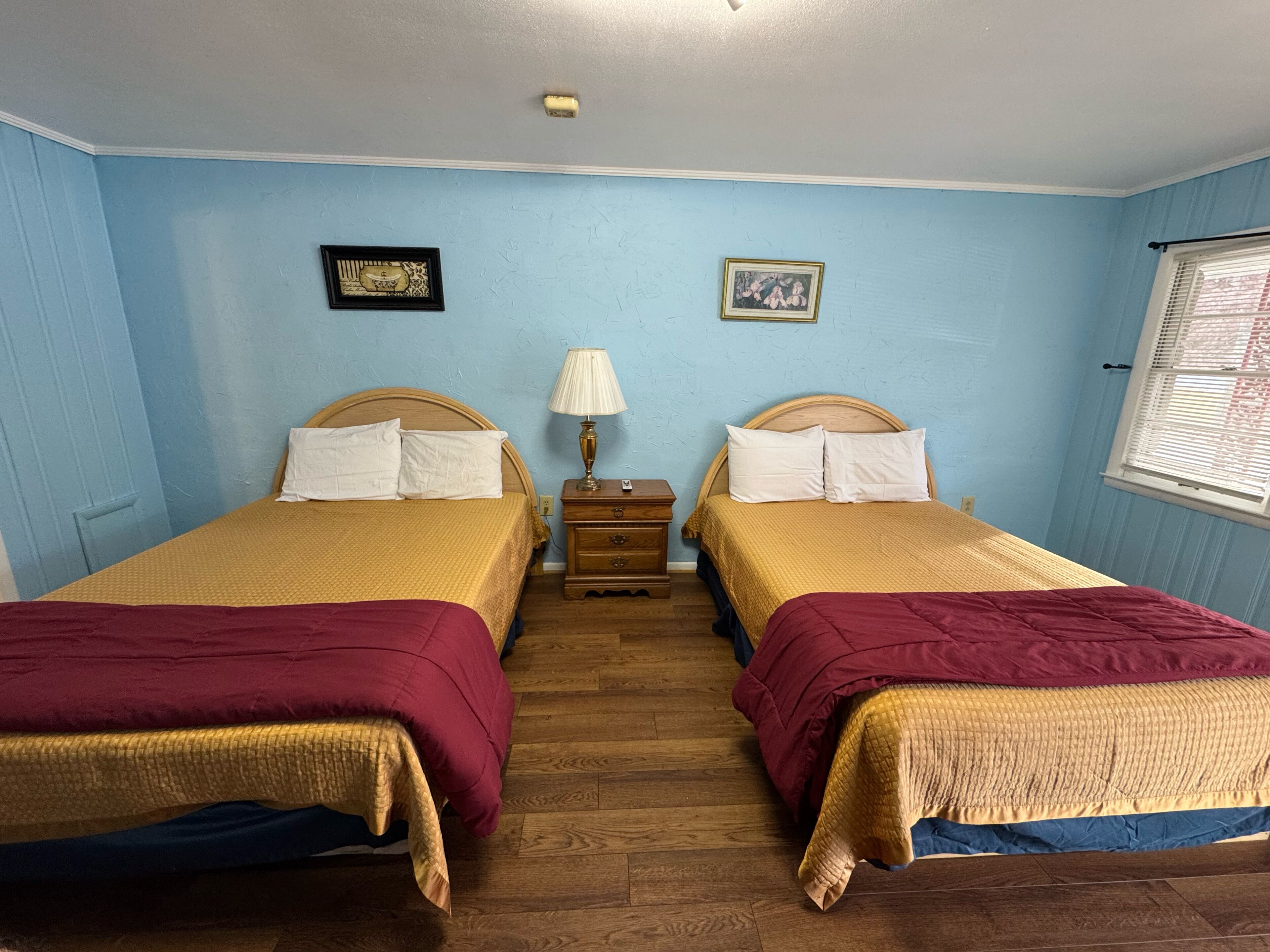 Ein Doppelzimmer mit zwei Betten, Holz-Kopfteilen, einem Nachttisch mit Lampe und zwei gerahmten Bildern an der Wand.