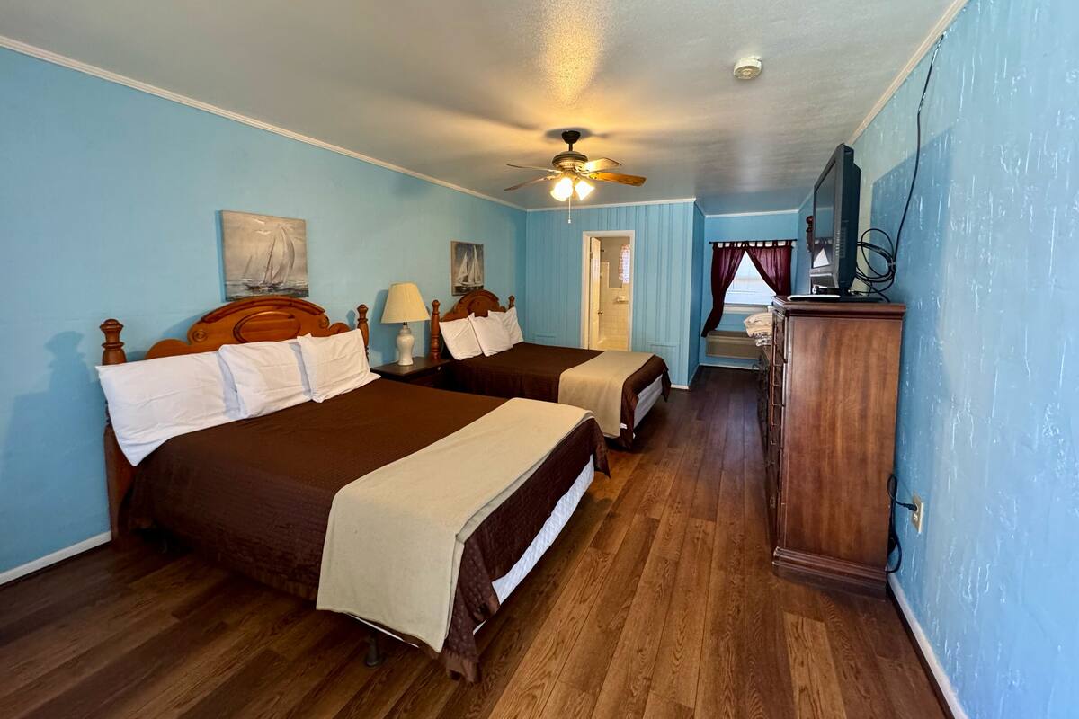 A room with two wooden beds, a ceiling fan, a wooden dresser, and a window with curtains.