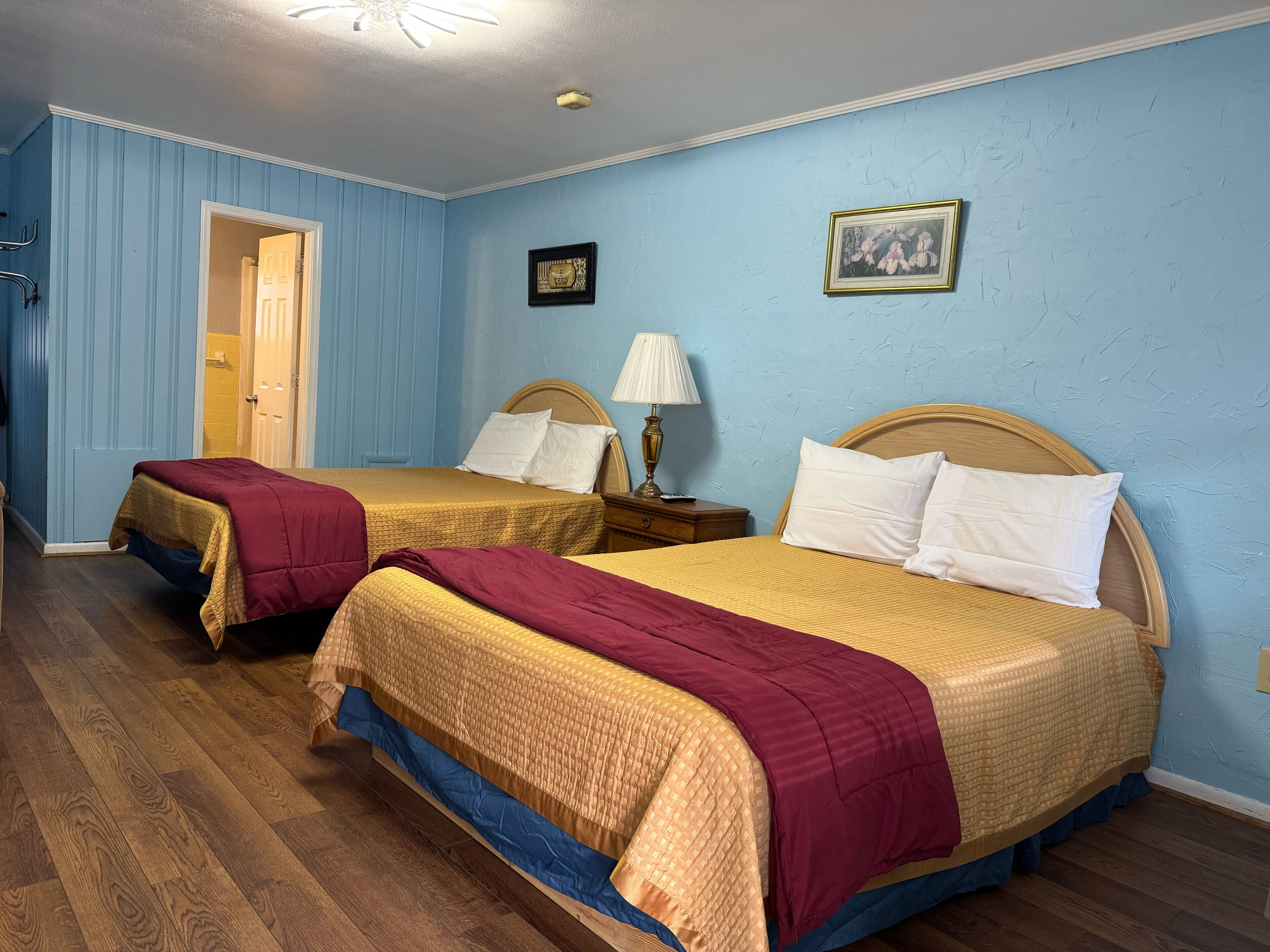 Habitación de hotel con dos camas, cabeceros de madera, una mesita de noche con lámpara y una puerta que da a otra habitación.