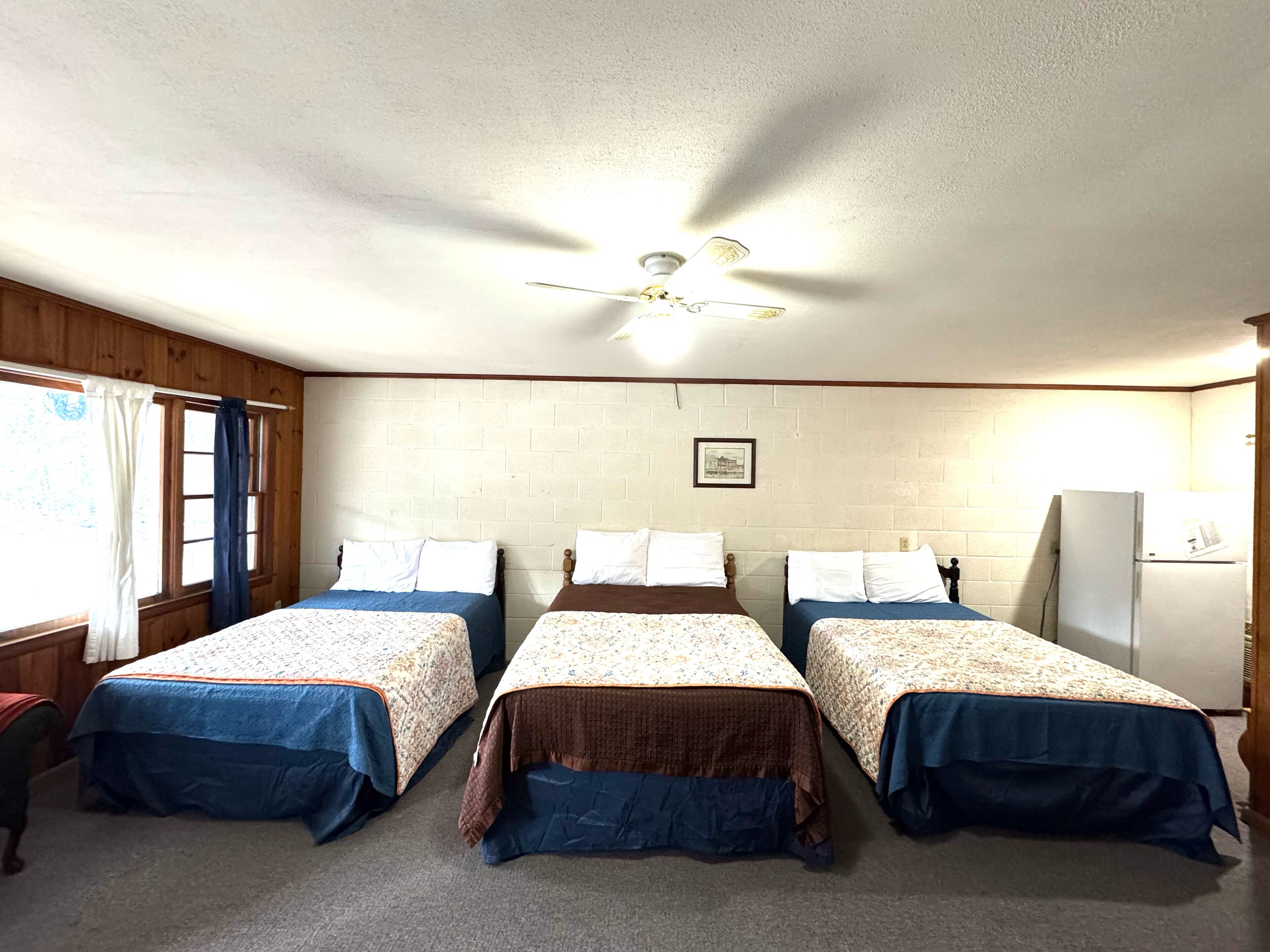 A room with three single beds, a ceiling fan, a window with curtains, and a refrigerator.