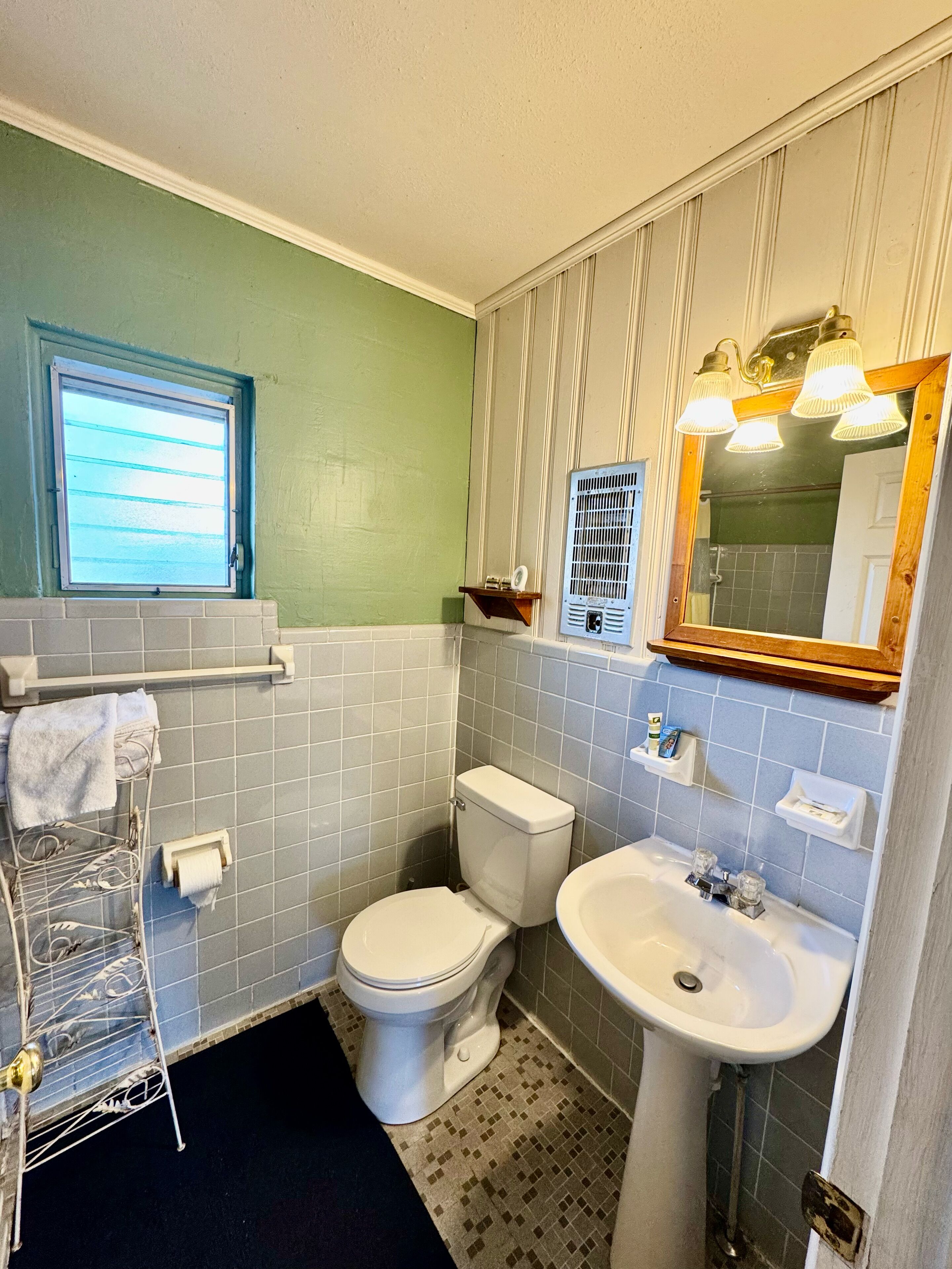 Un bagno con il water, un lavandino e uno specchio. Si vede una finestra sopra il water, e un porta asciugamani con degli asciugamani.