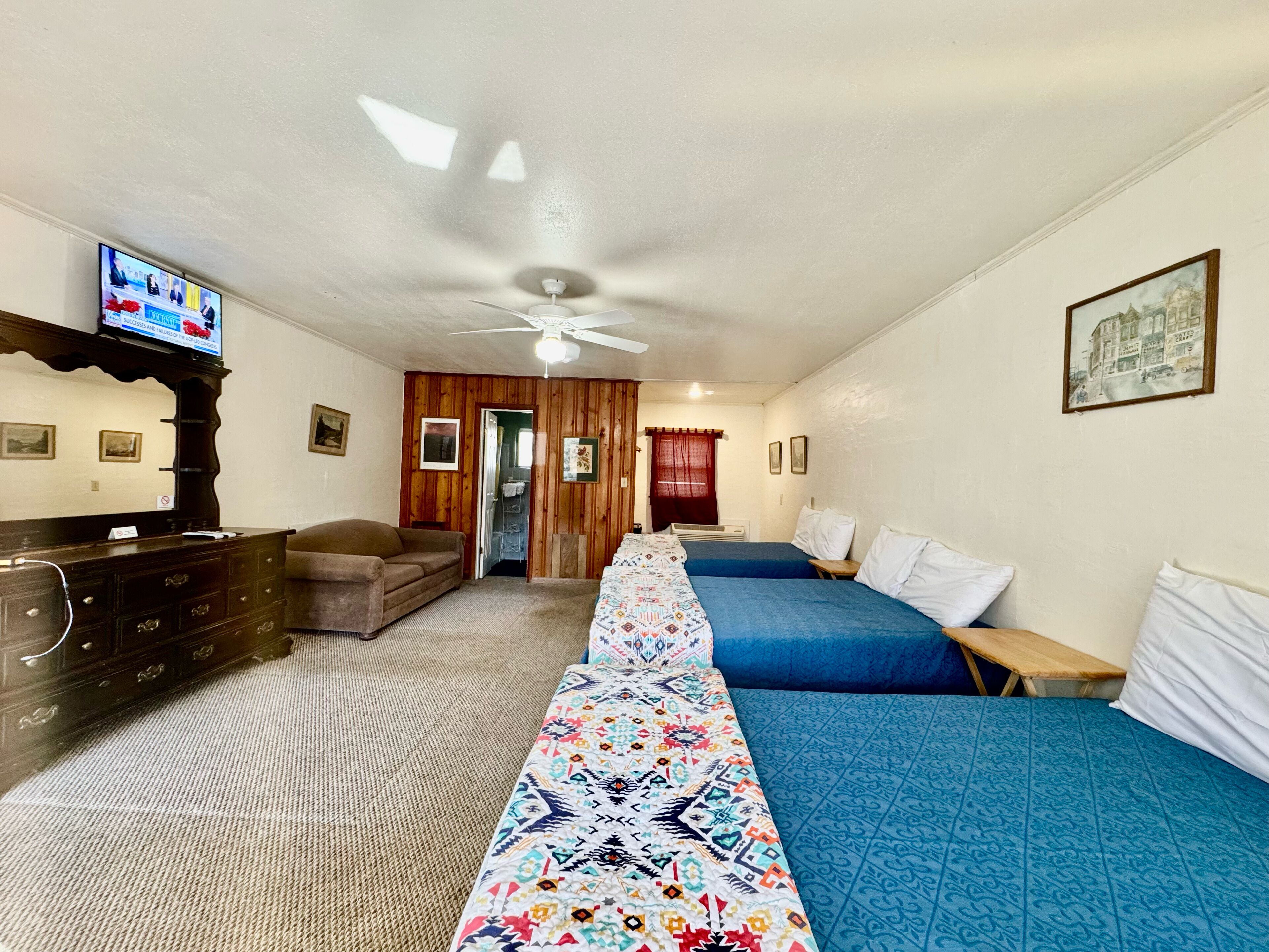 A hotel room with a long bed, two single beds, a TV, a dresser, and a ceiling fan.