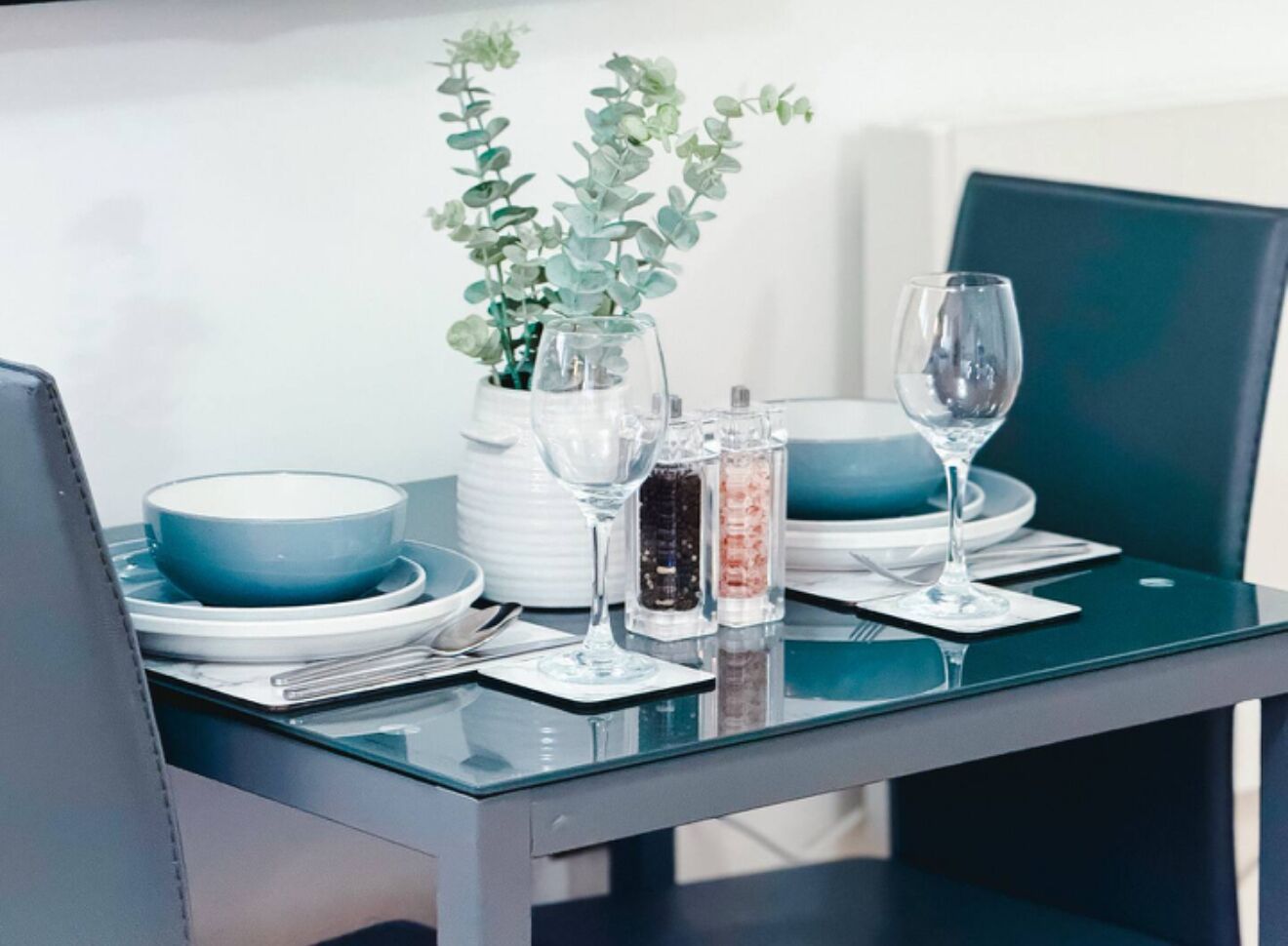 A dining table set with blue plates, clear glasses, and a vase with green plants.
