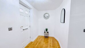 A hallway with white walls, a wooden floor, a door, a wall clock, a mirror, and a small side table with a plant. - Chic studio, sleeps 2, free parking (Pudsey)