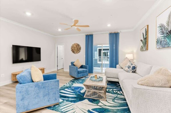 A living room with a flat-screen TV, a ceiling fan, and a large window with blue curtains.