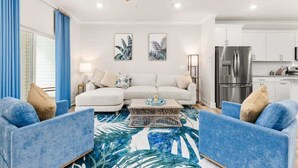 A living room with a sofa, two armchairs, a coffee table, and a ceiling fan. There are two framed pictures on the wall, a kitchen with a refrigerator, and a window with blue curtains.
