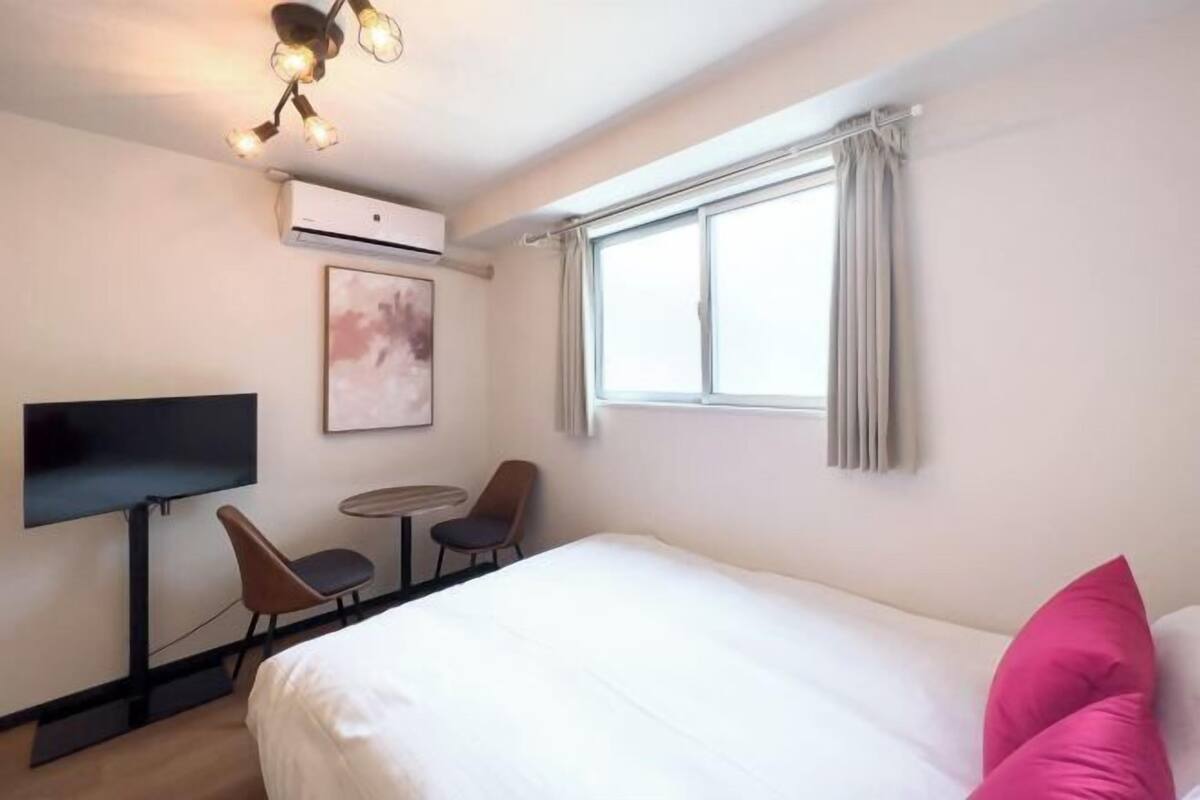 A hotel room with a bed, a TV, a small table, two chairs, an air conditioning unit, and a window with curtains.