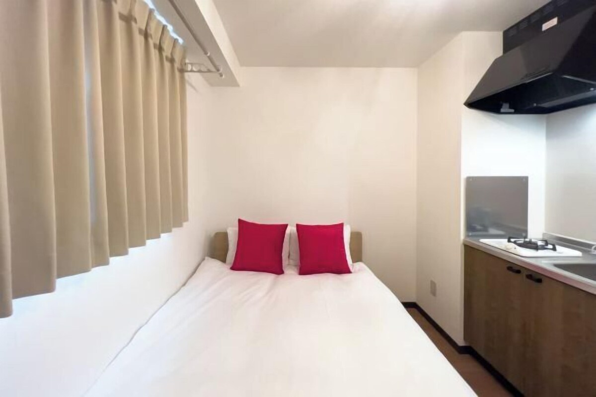 A hotel room with a bed, two red pillows, a window with blinds, a kitchen area with a stove, and a black range hood.