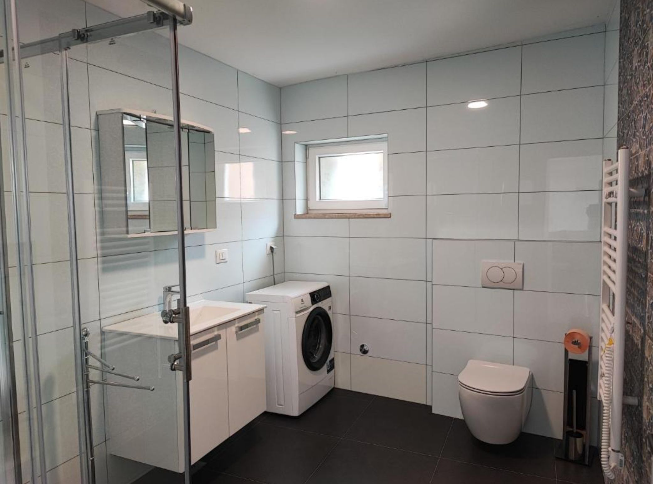 Ein kleines Badezimmer mit Duschkabine aus Glas, einer Waschmaschine, einer Toilette und einem kleinen Fenster für Tageslicht.