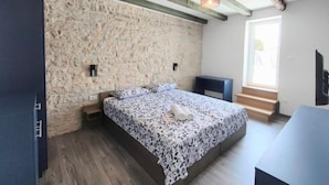 Une chambre avec un mur en pierre, un lit recouvert d’une couvre-lit à motifs floraux, un banc en bois et une fenêtre donnant sur un paysage agréable.
