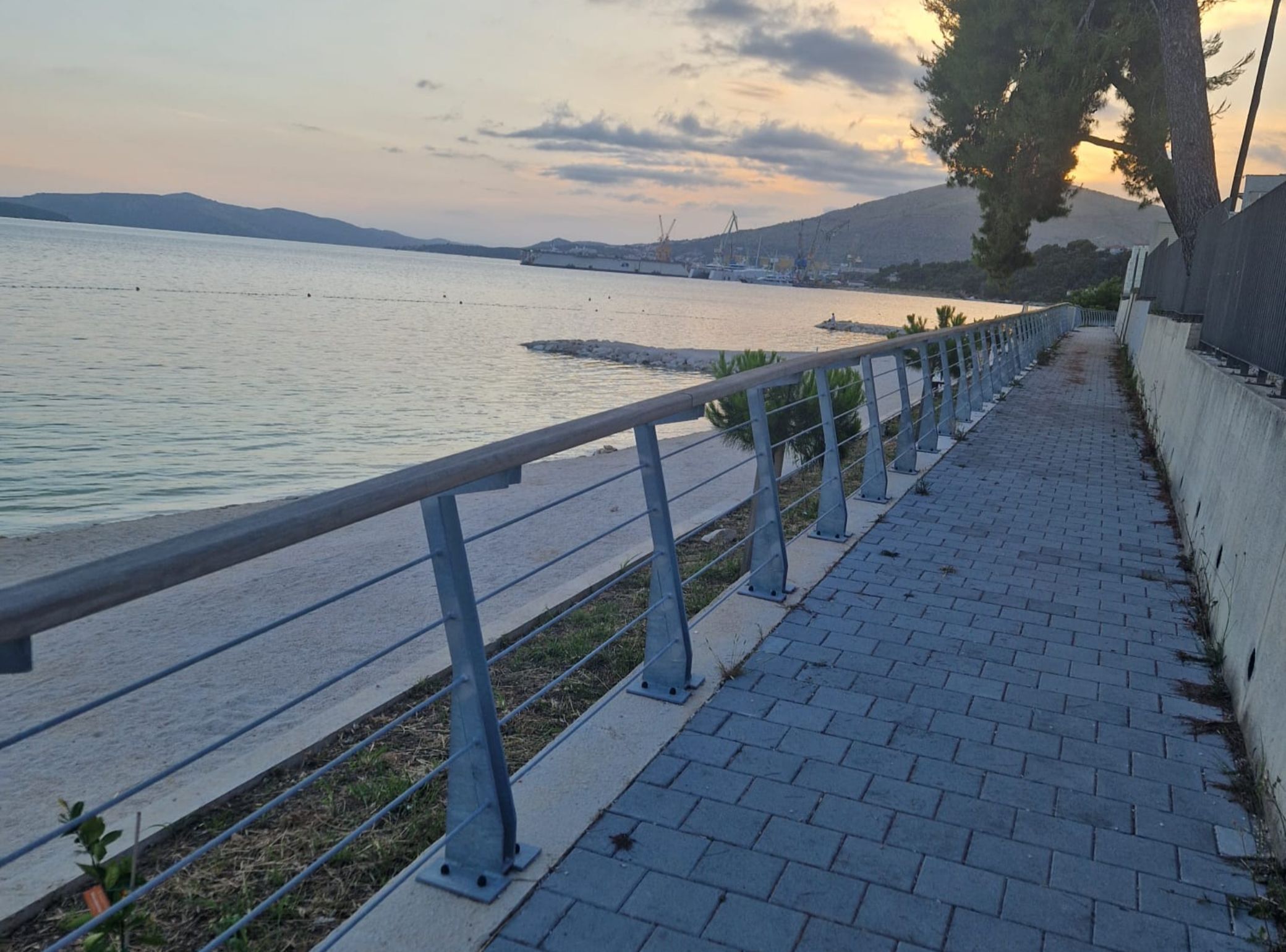 Een waterkantpromenade met een metalen hekwerk, een verharde looppad en bergen in de verte.