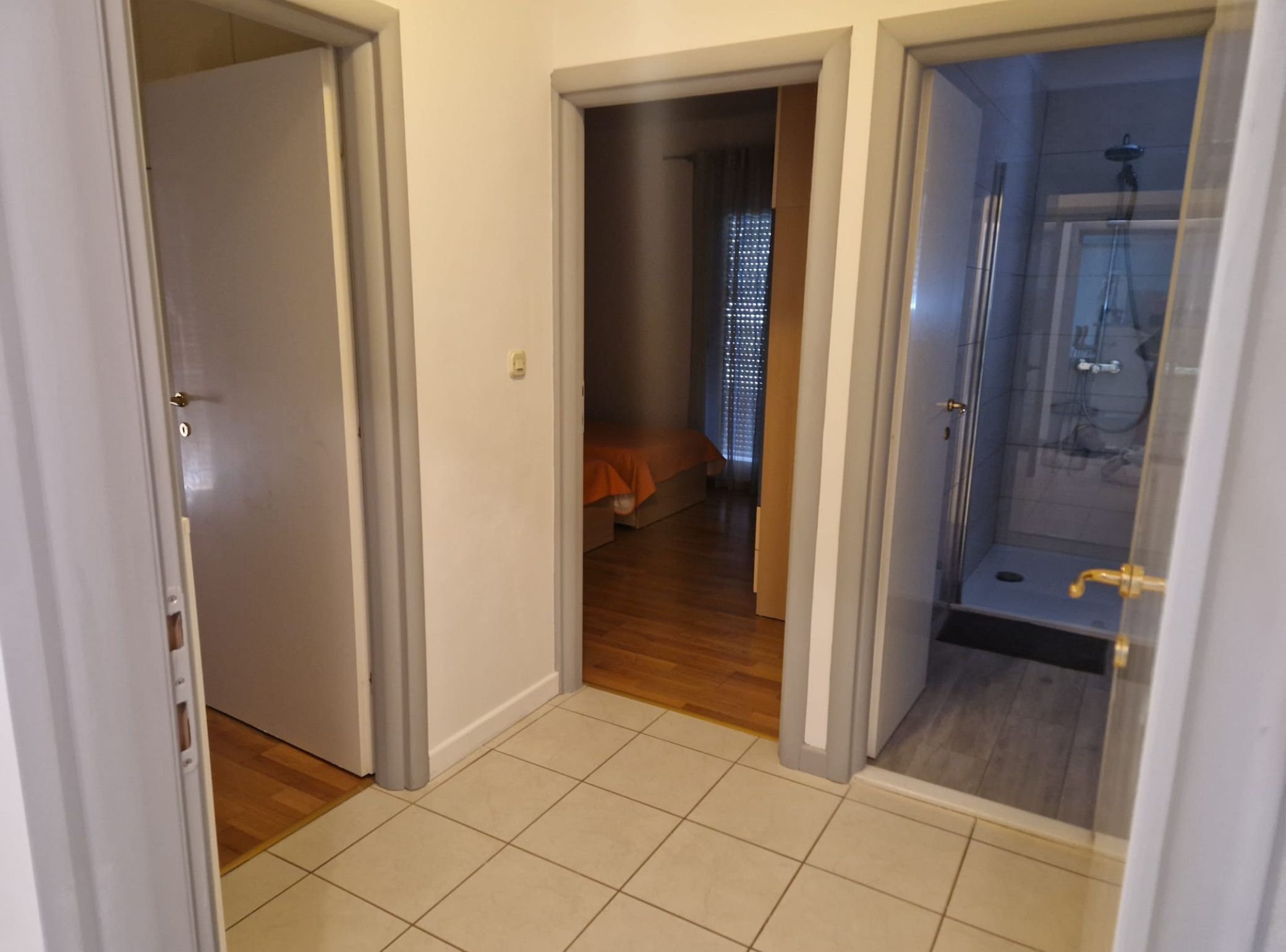 A hallway with a bathroom and a bedroom. The bathroom has a glass shower enclosure. The bedroom has a bed with an orange blanket.