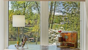 A room with a large window, a table with a lamp, and a wicker chair.