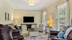A living room with a large flat-screen TV, leather sofas, a plaid rug, and a window with blinds.