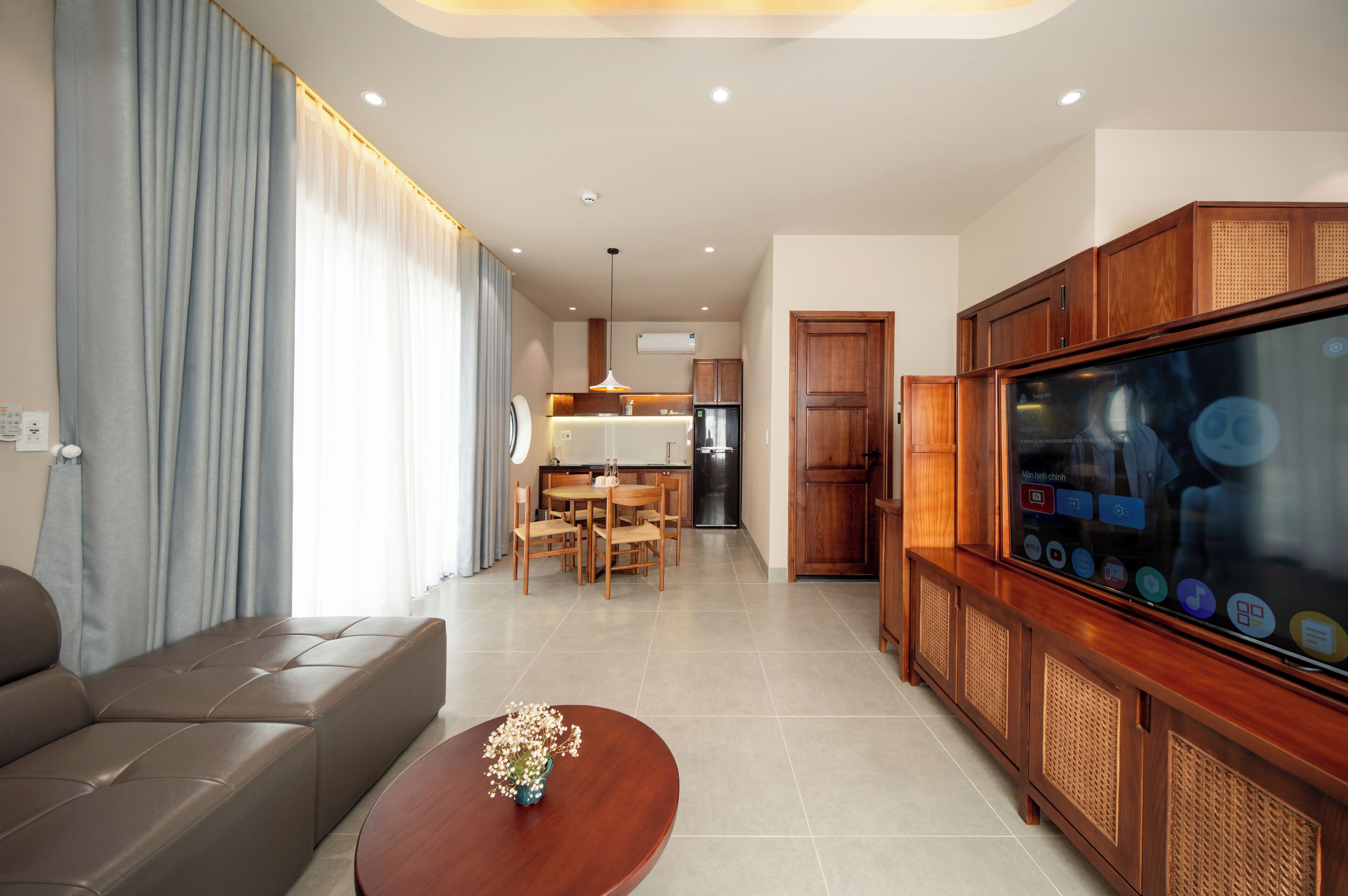 A modern living room with a sofa, a coffee table with flowers, a TV, and a kitchen in the background.