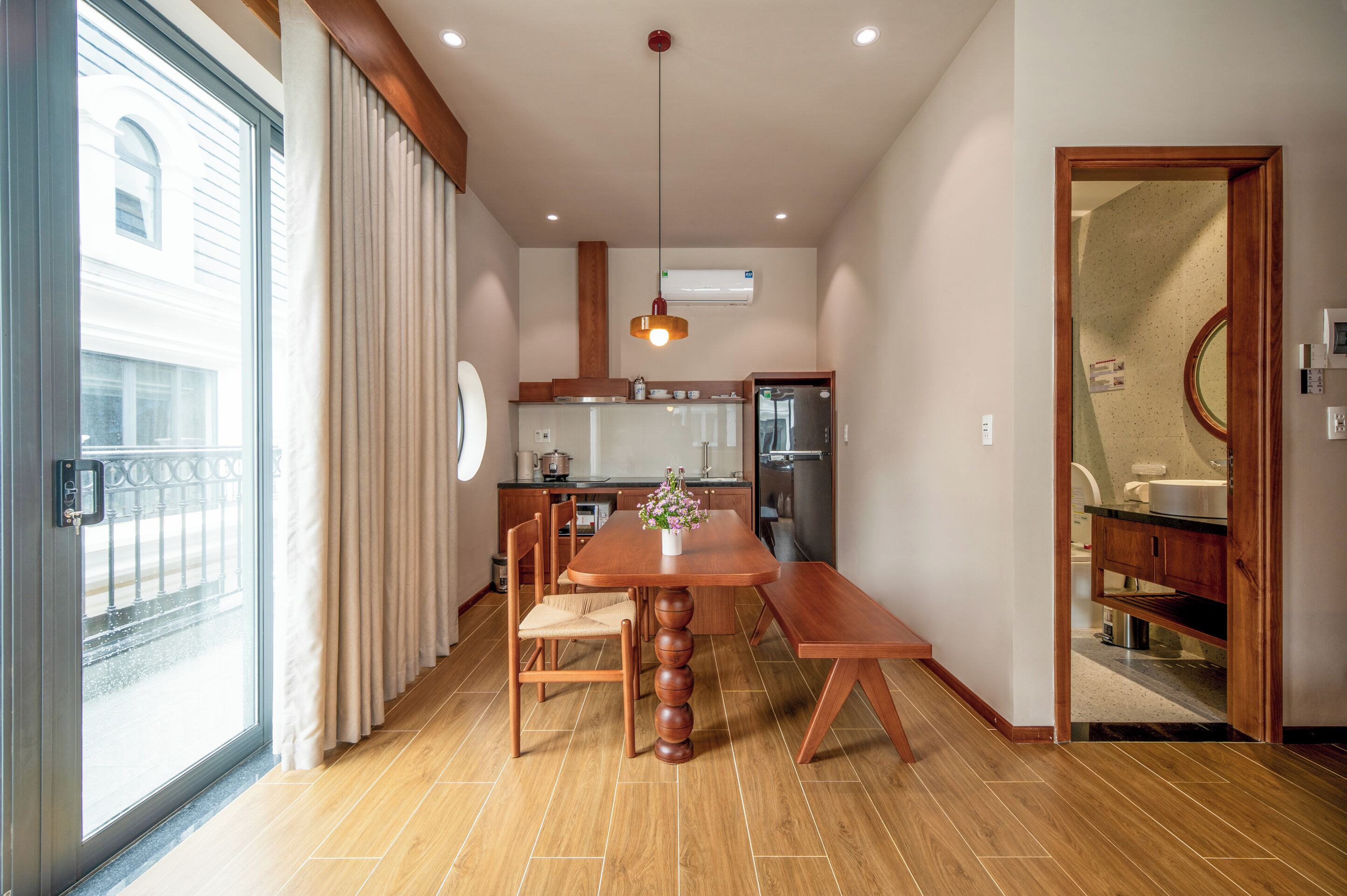 A modern kitchen with a dining area, wooden flooring, and a large window with curtains.