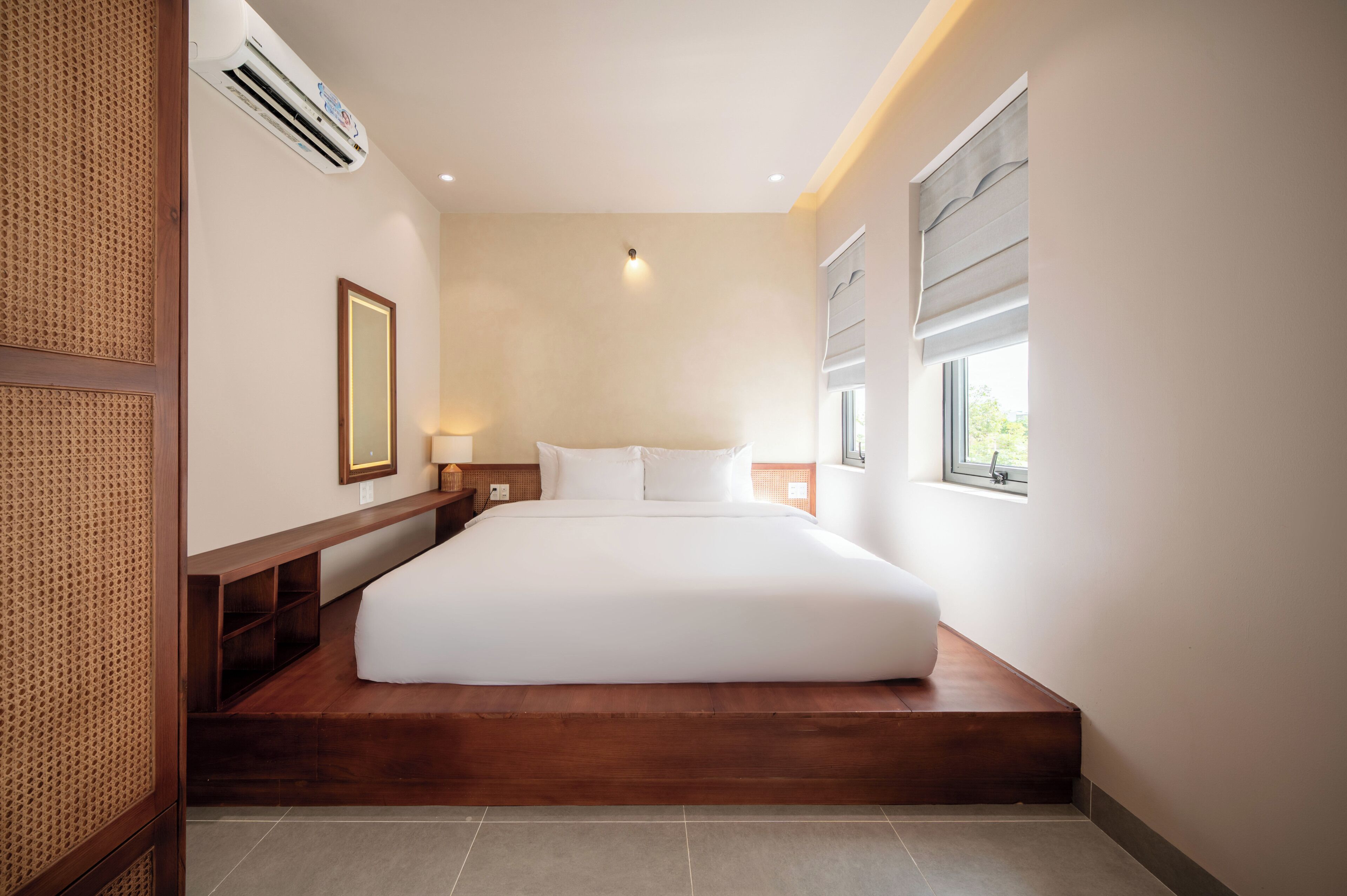 Quarto de hotel com cama, mesa de cabeceira, luminária, aparelho de ar condicionado, janela com persianas e moldura de quadro na parede.