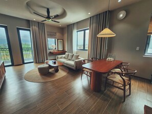 A modern living room with a sofa, coffee table, dining table, and chairs. There is a ceiling fan, large windows, and a view of the outdoors.