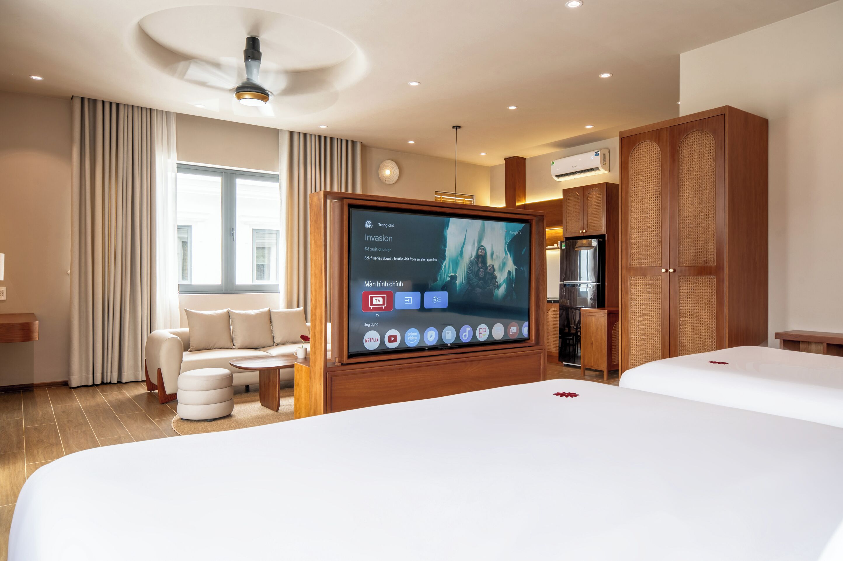 A hotel room with a large flat-screen TV, a sofa, a dining table, a minibar, and a wardrobe.
