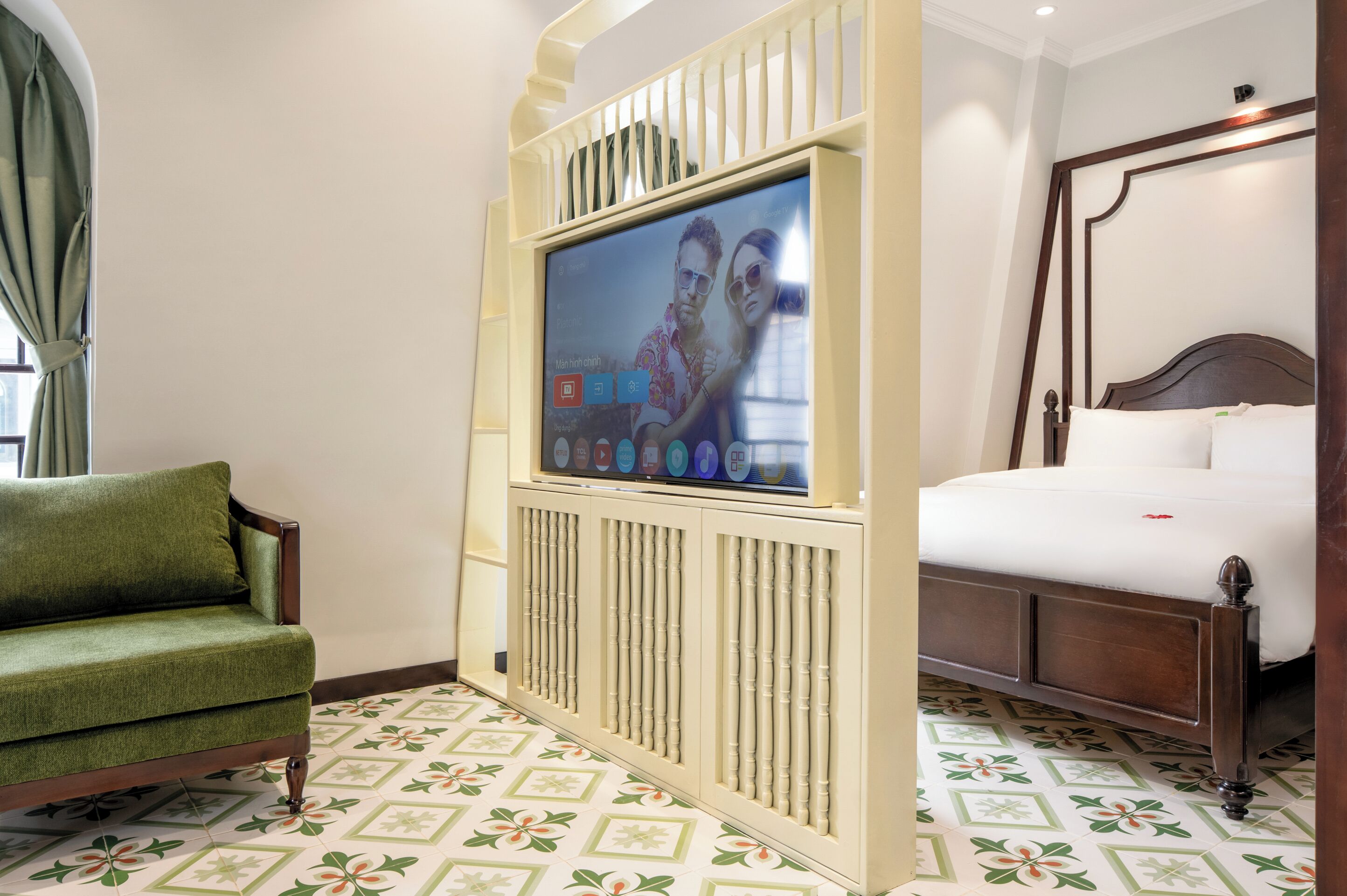 Quarto de hotel com cama, poltrona verde, televisão e piso com padrão.