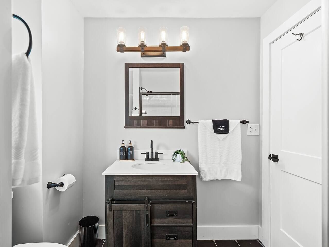 Une salle de bain avec un lavabo blanc, un meuble en bois foncé et un miroir surmonté d’appliques lumineuses.