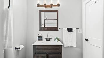 Une salle de bain avec un lavabo blanc, un meuble en bois foncé et un miroir surmonté d’appliques lumineuses.