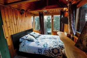Quarto de cabana de madeira com cama, mesa de cabeceira e janela com cortinas.