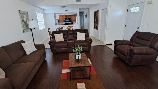 A living room with a brown leather sofa, a matching armchair, and a coffee table with a plant. A kitchen is visible in the background.