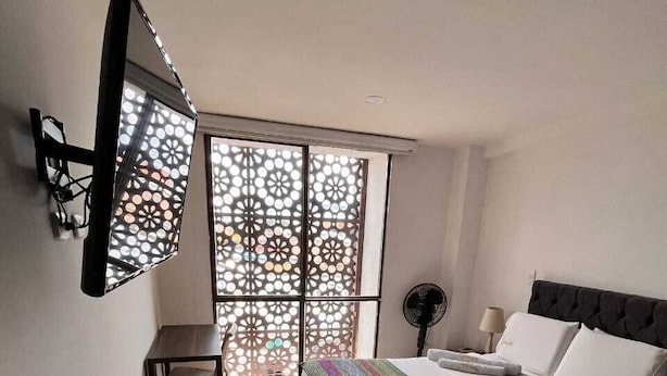 A bedroom with a bed, a desk, a chair, a standing fan, and a window with a decorative screen. - Samalá Casa Hotel Pitalito (Pitalito)