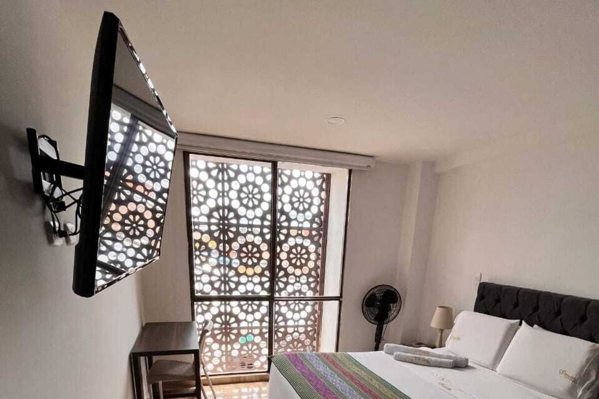 Une chambre à coucher comprenant un lit, un bureau, une chaise, un ventilateur sur pied et une fenêtre agrémentée d’un paravent décoratif.