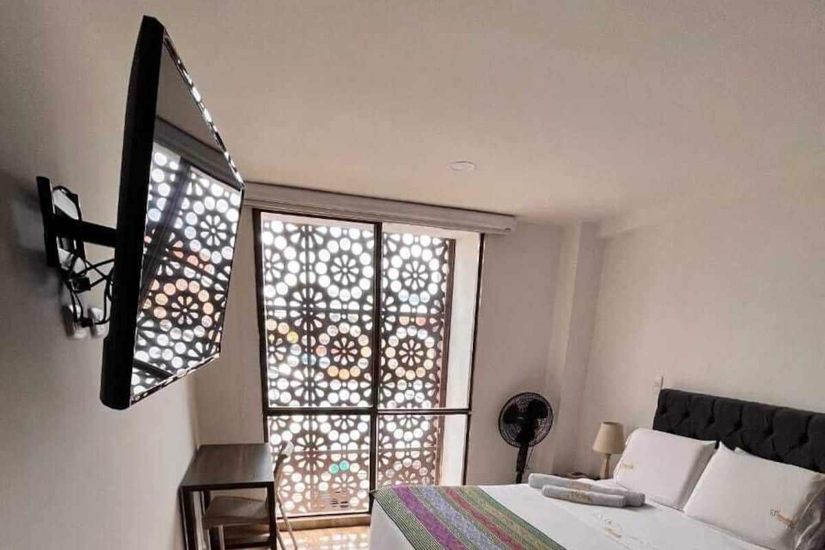 A bedroom with a bed, a desk, a chair, a standing fan, and a window with a decorative screen.