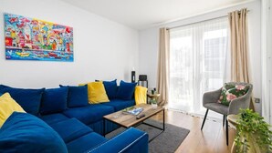 Una sala de estar con un sofá azul, cojines amarillos, una mesa de centro, una silla, un cuadro colorido y un ventanal con cortinas.
