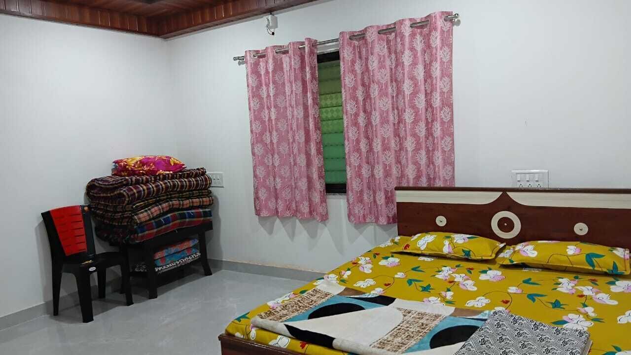 A bedroom with a bed, a chair, a small table with folded blankets, and a window with pink curtains.