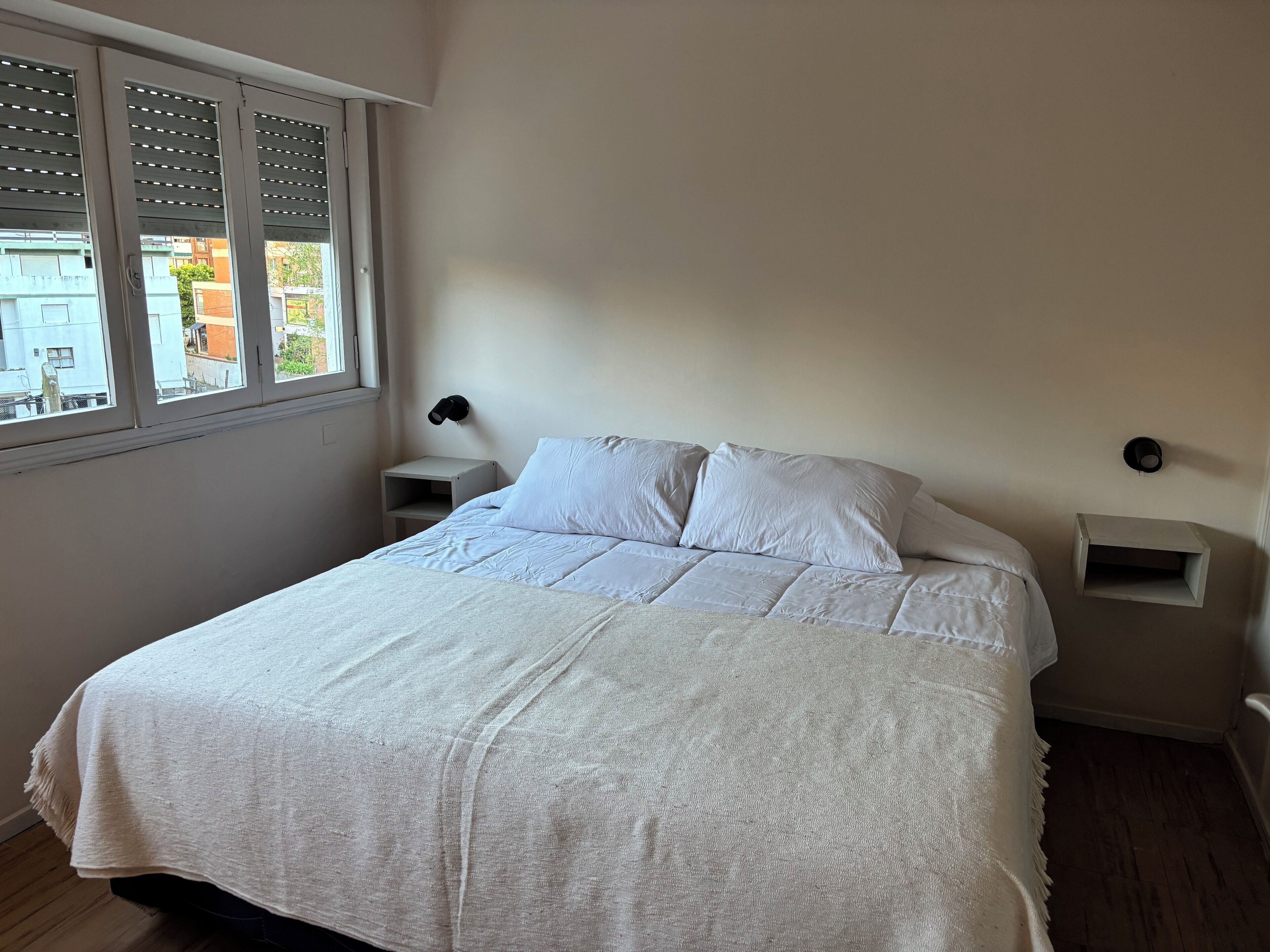 A bedroom with a large bed, two bedside tables, a window with blinds, and a view of buildings outside.