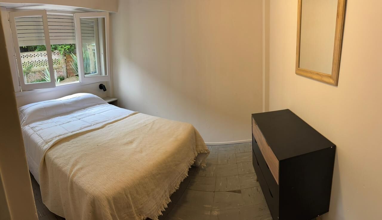 A bedroom with a bed, a bedside table, a small black cabinet, and a window with shutters.