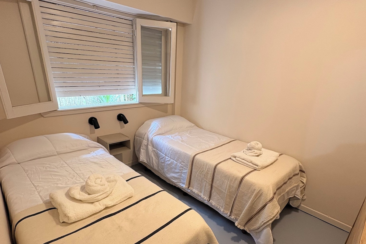 A small, compact room with two single beds, each with a striped blanket and a rolled towel. There is a window with blinds, and the walls are painted a light color.