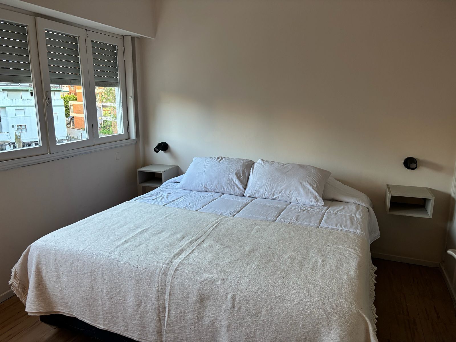 A bedroom with a bed, two bedside tables, a window, and a view of buildings outside.