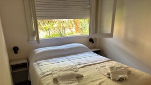 A neatly made bed with a white duvet and two folded towels, a bedside table with a lamp, and a window with blinds. - Black Suites Pinamar  (Pinamar)