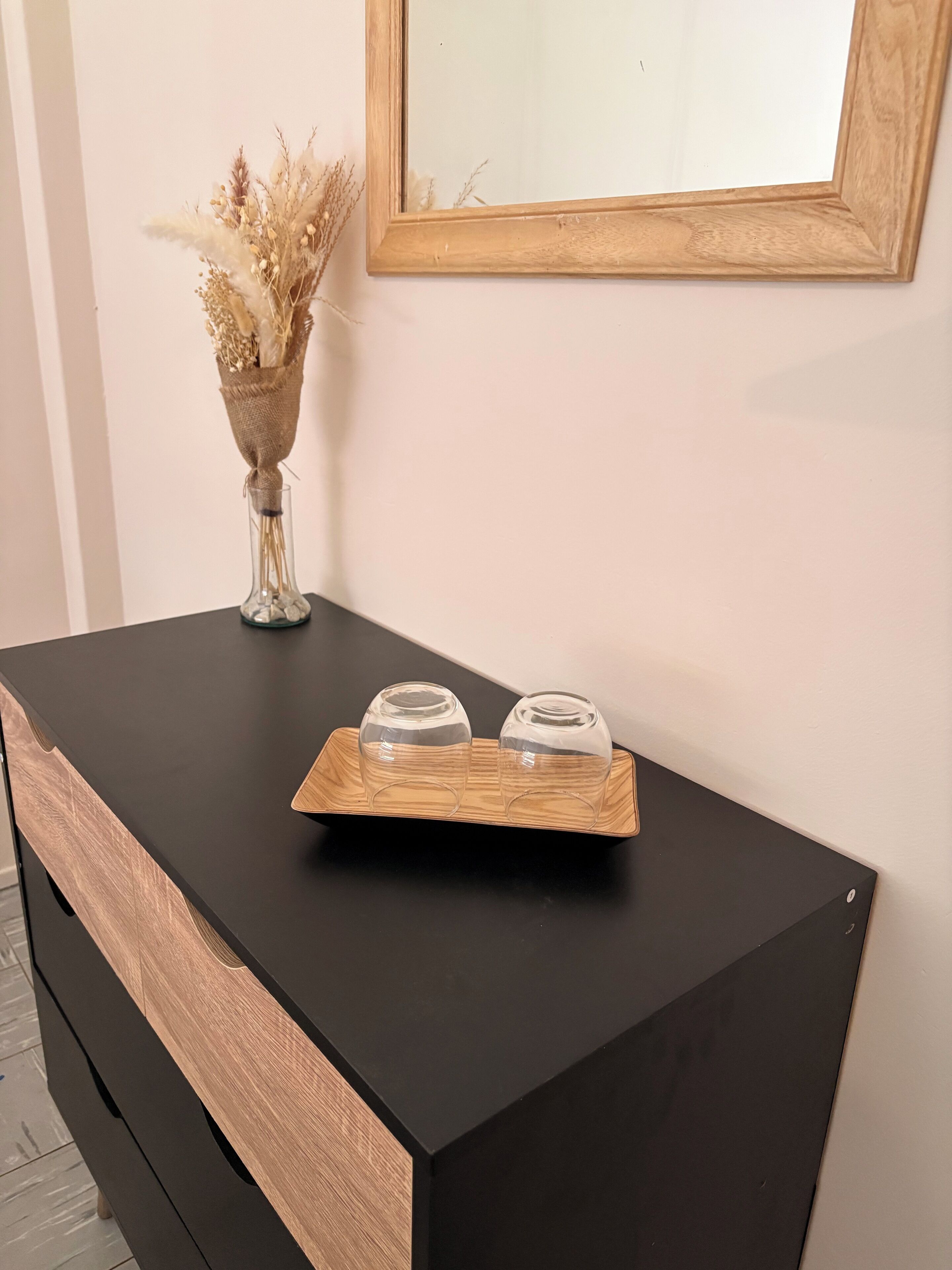 A black console table with a wooden tray holding two clear glass containers. A vase with dried flowers is placed on the wall behind the table.