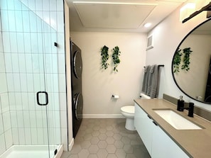 A modern bathroom with a white tiled shower, a toilet, a sink with a mirror, and green plants on the wall.