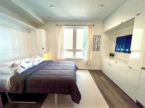 Un dormitorio con cama, televisor, ventana con cortinas y un mueble de pared.