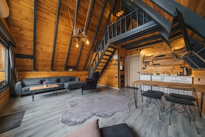 Un loft spacieux avec de hauts plafonds, des poutres apparentes, un escalier, un coin salon avec un canapé et une table basse, et un espace cuisine.