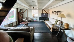Una sala de estar acogedora con sofá, sillón y televisor. Hay una estantería, una lámpara y una ventana con vista a un vehículo verde afuera.
