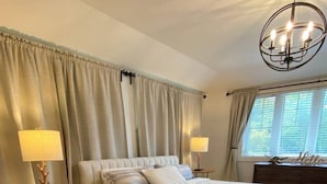 A bedroom with a large bed, two bedside lamps, a chandelier, a window with curtains, and a rug. - Private Beach, Hot Tub, A Vacay to Remember! (Prince Edward)