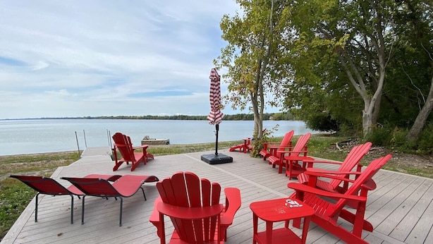 A wooden deck with red Adirondack chairs and tables, overlooking a body of water. - Private Beach, Hot Tub, A Vacay to Remember! (Prince Edward)